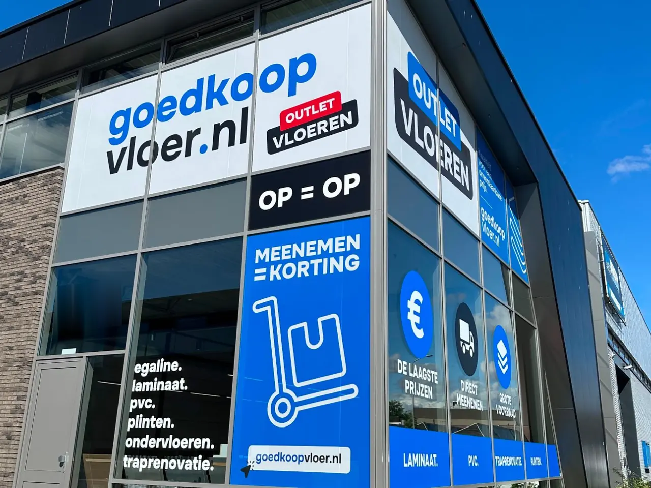 Hoek van de winkel van Goedkoopvloer met het logo over drie ruiten, een outlet-sticker, een blauw raamvlak en een strook met productgroepen